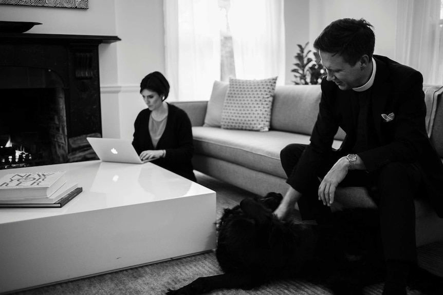 A woman sits at a laptop by a fire and a man sits near her on a couch with a dog