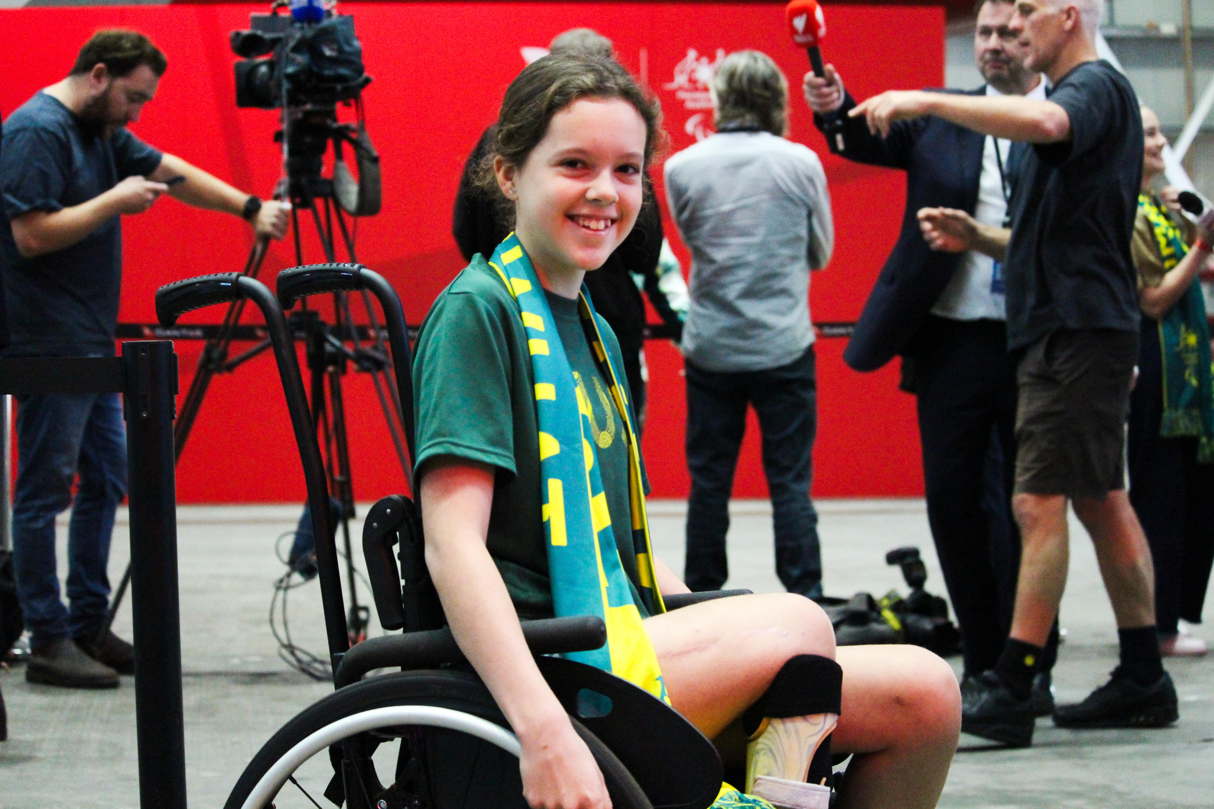 A young girl in a wheelchair, wearing a green shirt and green and gold scarf, smiling