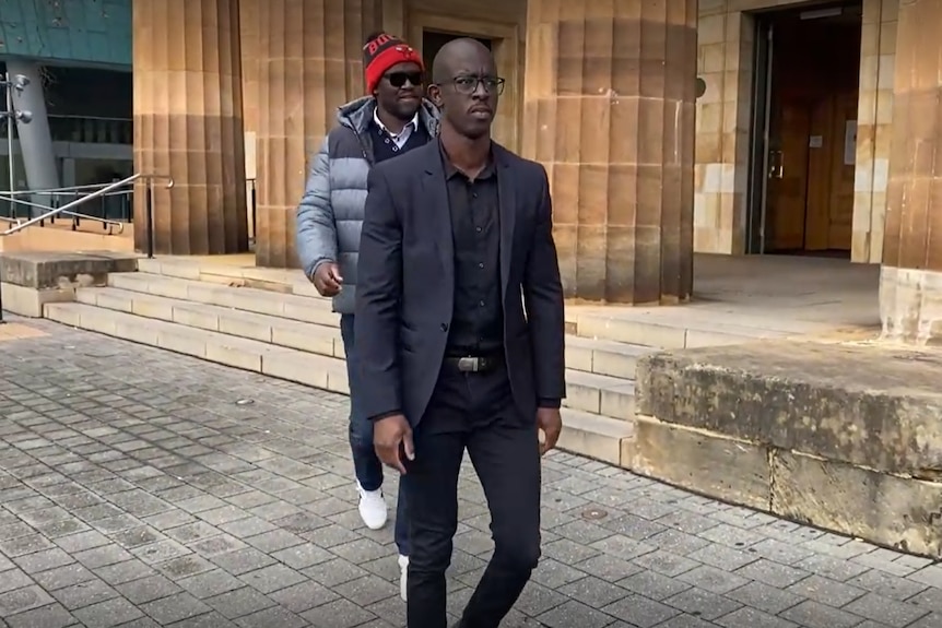 Martin Duku walking outside a court building wearing a suit and glasses