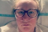 A woman wearing glasses lays in a hospital bed with a tube inserted into her nostrils.