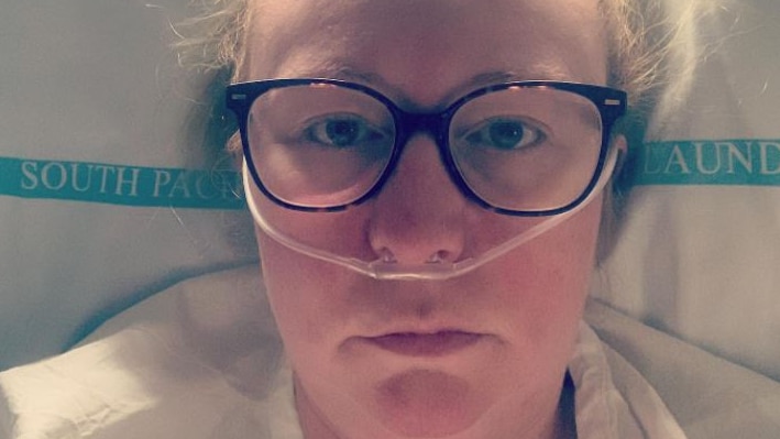 A woman wearing glasses lays in a hospital bed with a tube inserted into her nostrils.