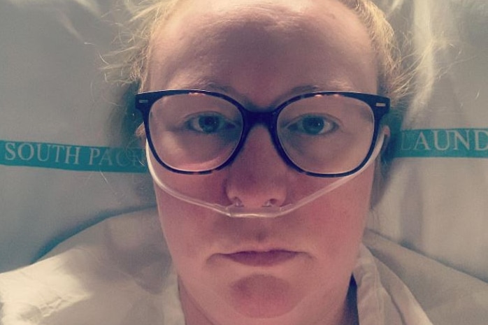 A woman wearing glasses lays in a hospital bed with a tube inserted into her nostrils.
