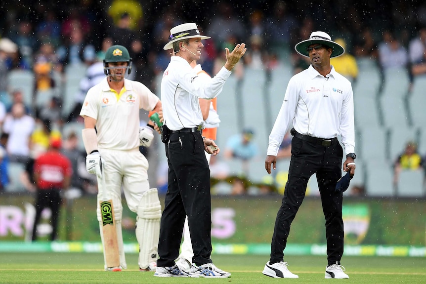 A man raises his palms to the sky as rain falls, with a batsman and another man looking on