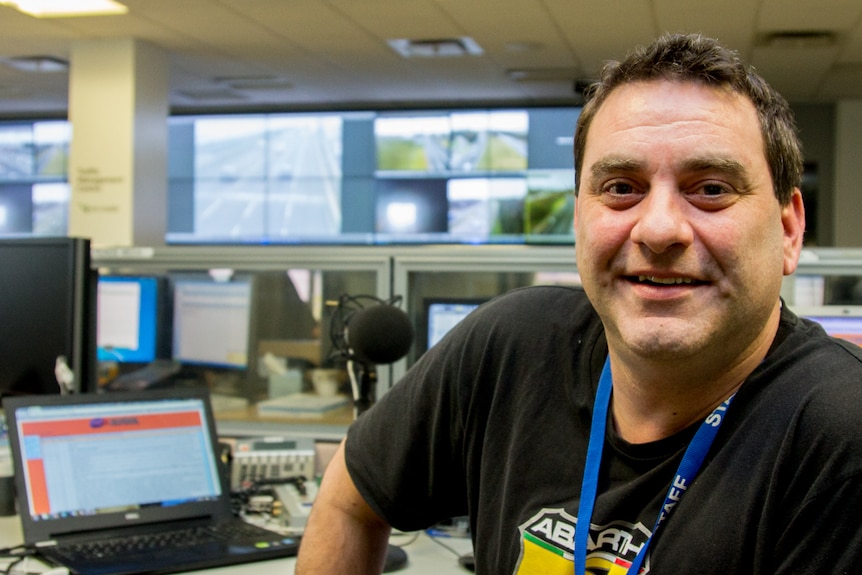 A man at a desk with a microphone, video wall showing freeways in background.