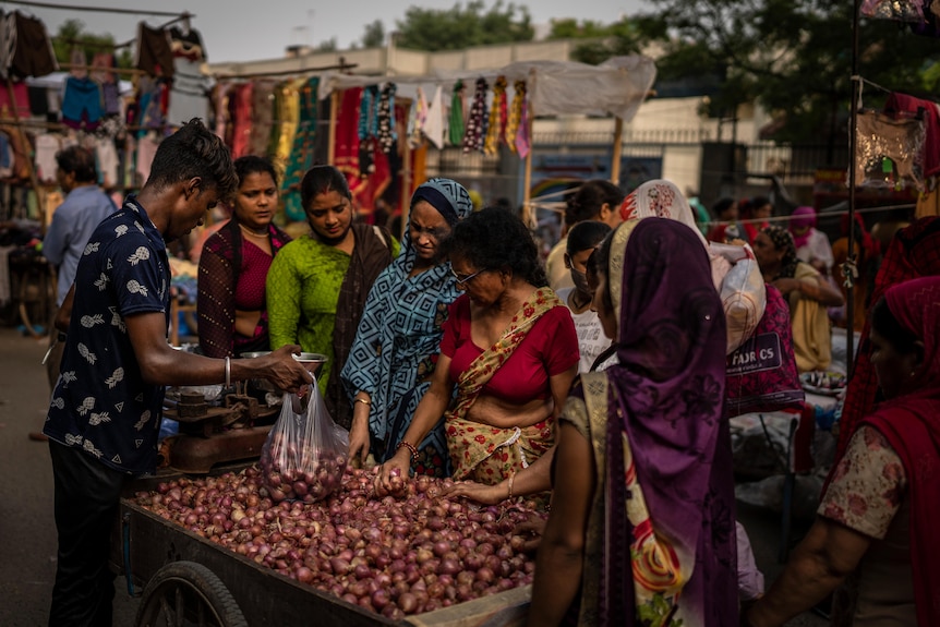 A roadside vendor packs red onions in plastic bags as women shop at a weekly market.