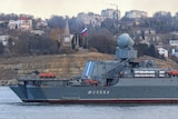 A large military ship with the name Moskva written across it sits in water near land where a Russian flag stands.
