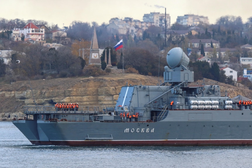 A large military ship with the name Moskva written across it sits in water near land where a Russian flag stands.