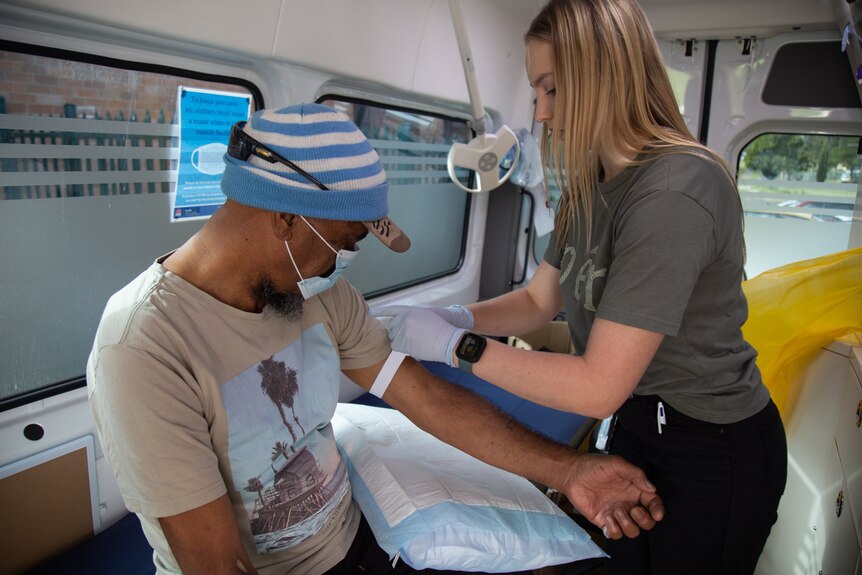a man wearing a cap getting a needle put into his upper arm by a woman wearing plastic gloves on her hands