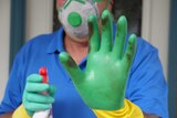 A man standing at his front door wearing cleaning gloves holds and holding a spray bottle holds his hand up.