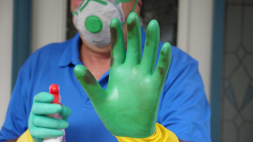 A man standing at his front door wearing cleaning gloves holds and holding a spray bottle holds his hand up.