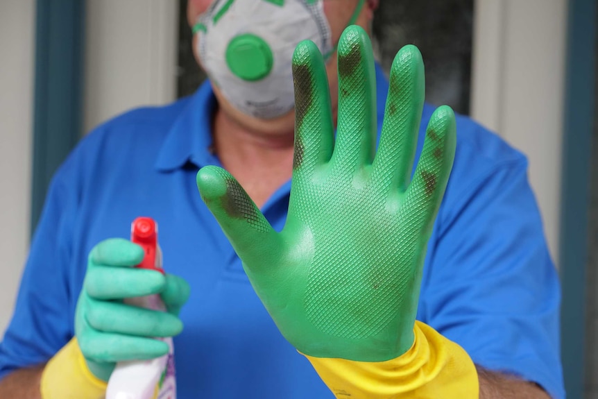 A man standing at his front door wearing cleaning gloves holds and holding a spray bottle holds his hand up.