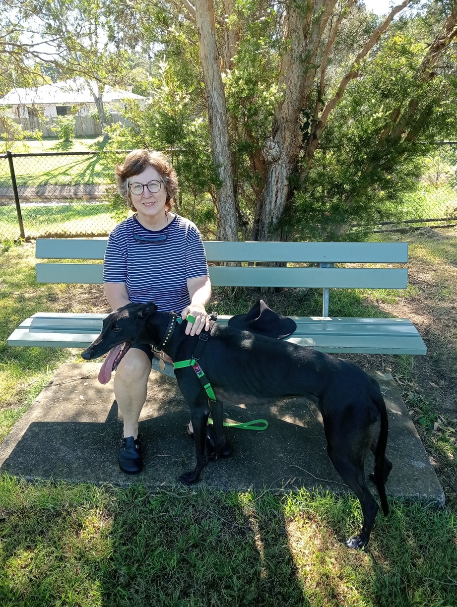 A smiling woman sitting on a wood park bench, patting a large black greyhound.