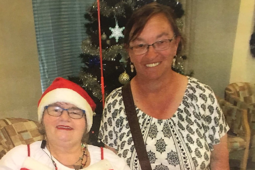 Debbie sits next to a friend in a red outfit in front of a Christmas tree.