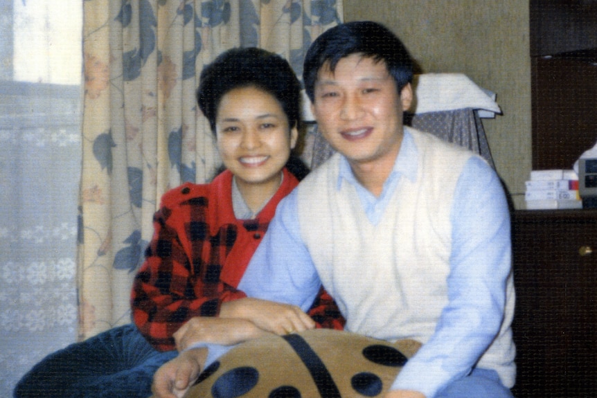 Young Xi Jinping, wearing a white jacket over a blue shirt, cuddles with Peng Liyuan in a red sweater