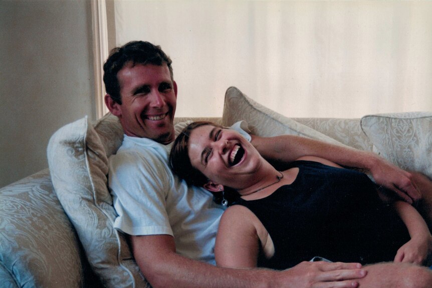 A man and woman on the couch. He is smiling while she is laughing.