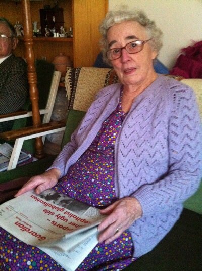 Beryl Miller, wearing purple cardigan and dress, sits on a chair holding a newspaper on her lap, with neutral expression.