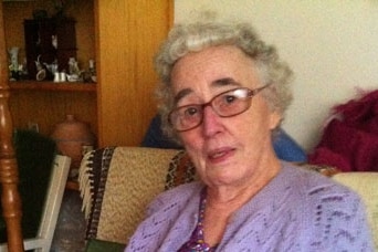 Beryl Miller, wearing purple cardigan and dress, sits on a chair holding a newspaper on her lap, with neutral expression.