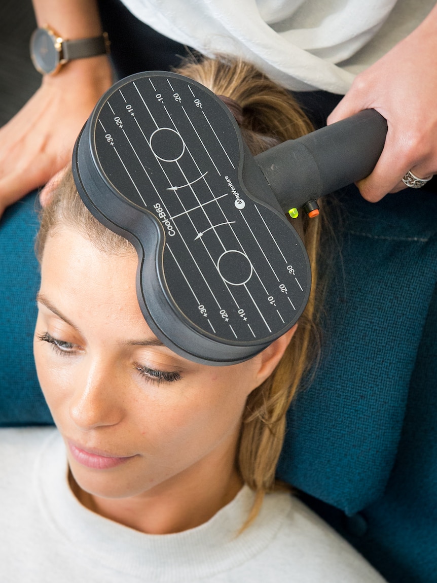 A woman sits in a chair while a figure 8 shaped scanner is held to their head by another person
