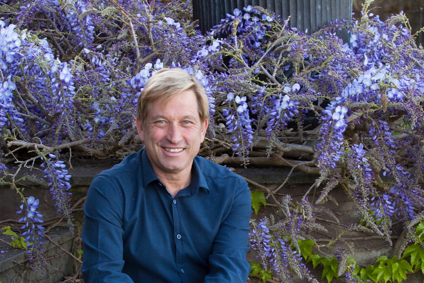 Paul is sitting on some stairs wearing a navy blue shirt and khaki pants. He has blond hair and blue eyes.