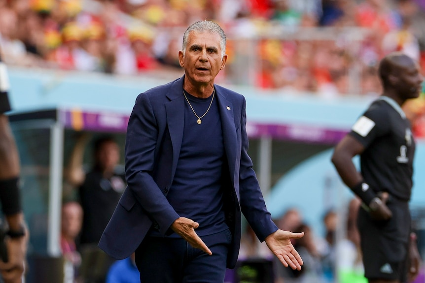 An Iranian football coach wearing a blue suit stands on the touchline gesturing with his hands during a game. 