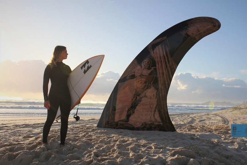 Female surfer looking at sculpture of large surfboard fin on a beach.