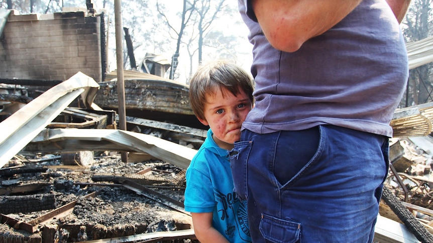 A child clinging to a parent gives a solemn look to the camera against the backdrop of his burnt and ruined home