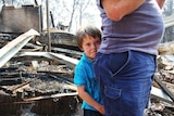 A child clinging to a parent gives a solemn look to the camera against the backdrop of his burnt and ruined home