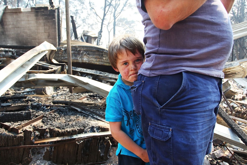 A child clinging to a parent gives a solemn look to the camera against the backdrop of his burnt and ruined home
