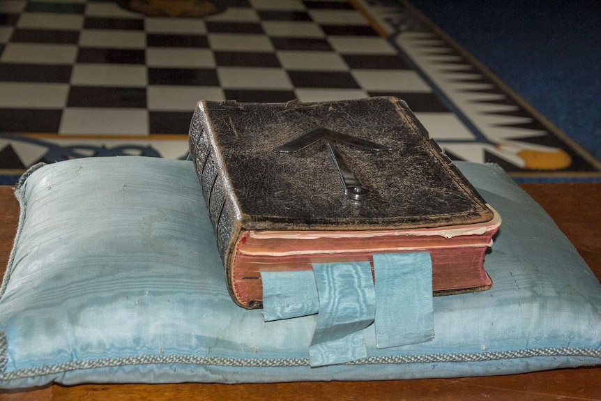 Bible with masonic symbols of architectual tools of the square and compass sitting on faded satin pillow