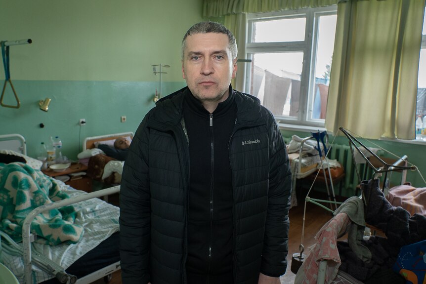 A man in a black puffer looking stern in a hospital ward 