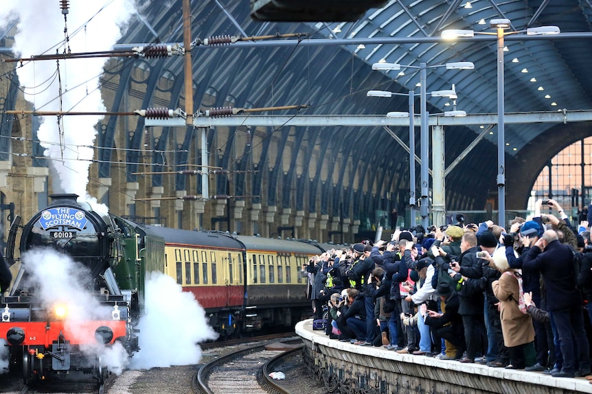 The Flying Scotsman, steam billowing, leaves Kings Cross station as a crowd of people on a platform opposite look on.