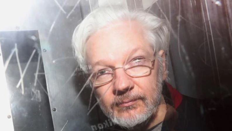 A photograph of Julian Assange through the window of what appears to be a police van.