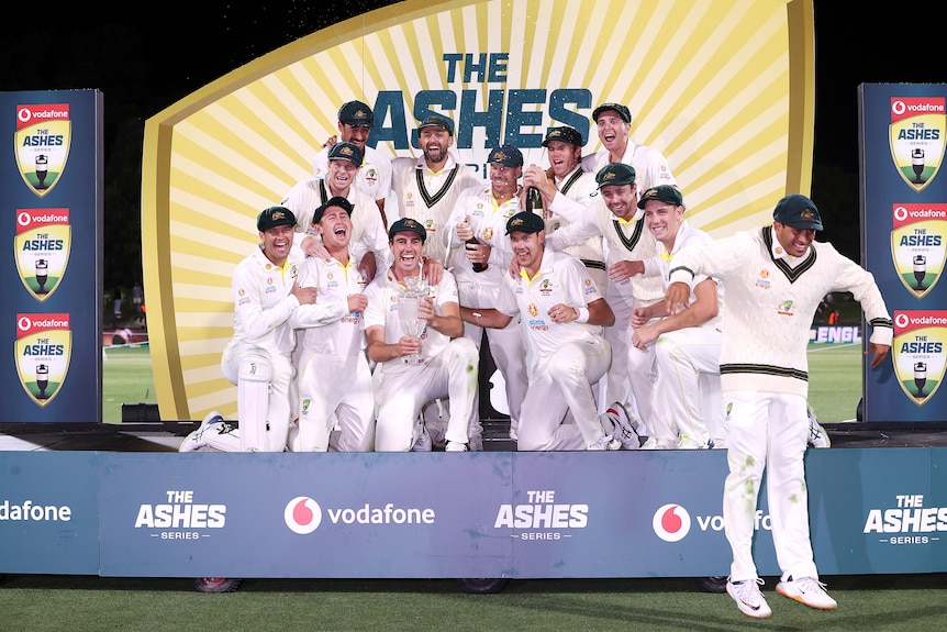 A group of Australian cricketers celebrate and start to pop champagne, as one player jumps off the stage to avoid the spray.