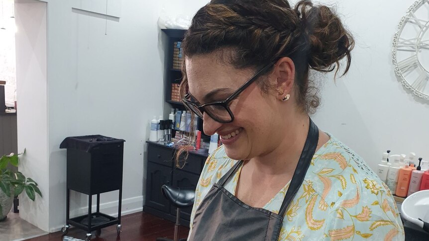 A woman wearing an apron in a hair salon looking down at her client's head and smiling.