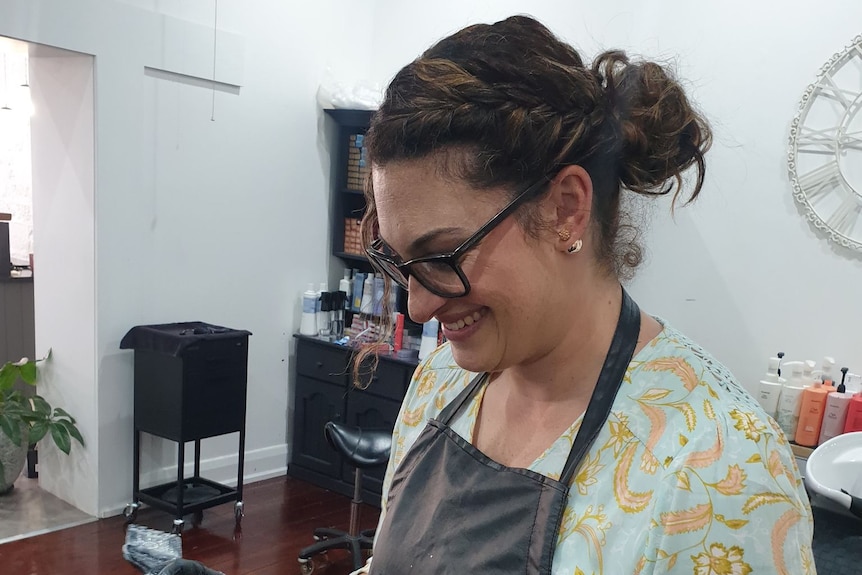 A woman wearing an apron in a hair salon looking down at her client's head and smiling.
