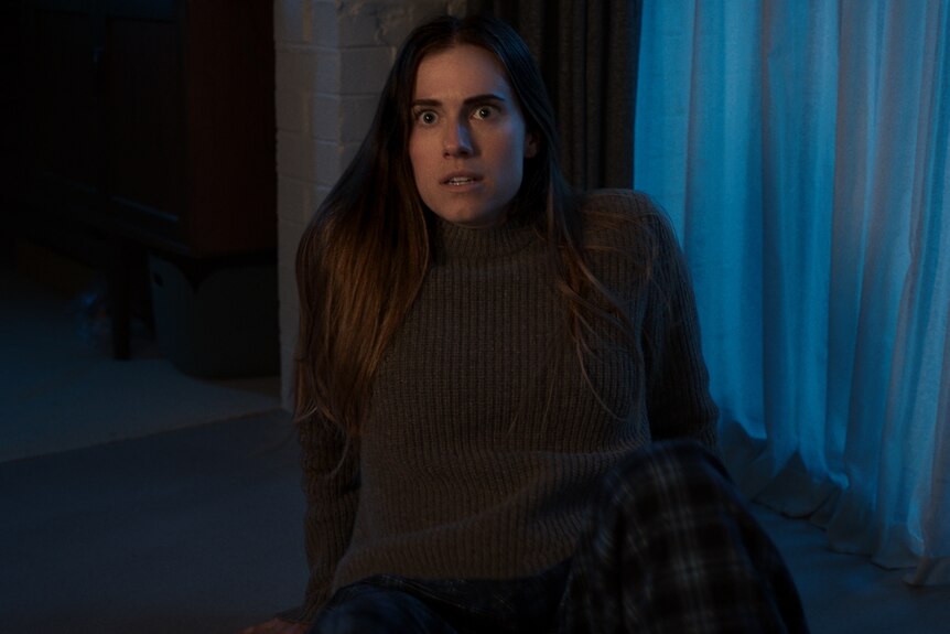 A white woman with long mousey brown hair wears an olive jumper and recoils on the floor of a living room.