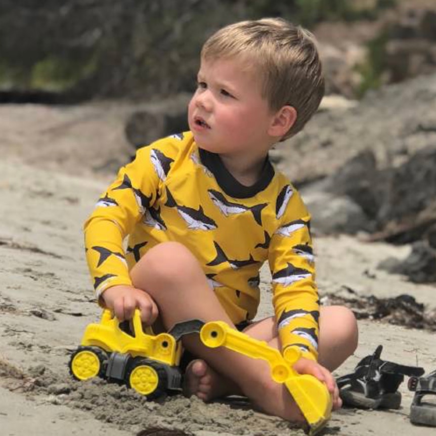 A two-year-old boy sits in sand, playing with a toy tractor.