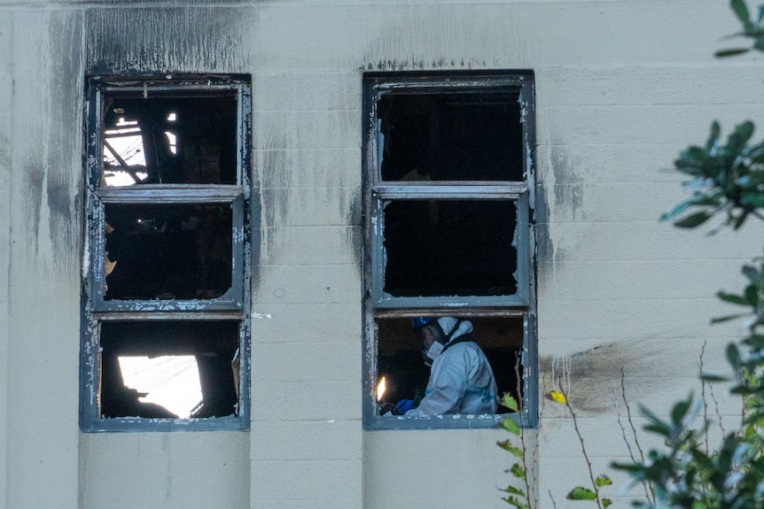 A person wearing protective gear including face mask and full-body suit is seen through the scorched window of a building
