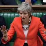 Theresa May addresses Parliament in a bright reddish orange jacket, with both hands up.