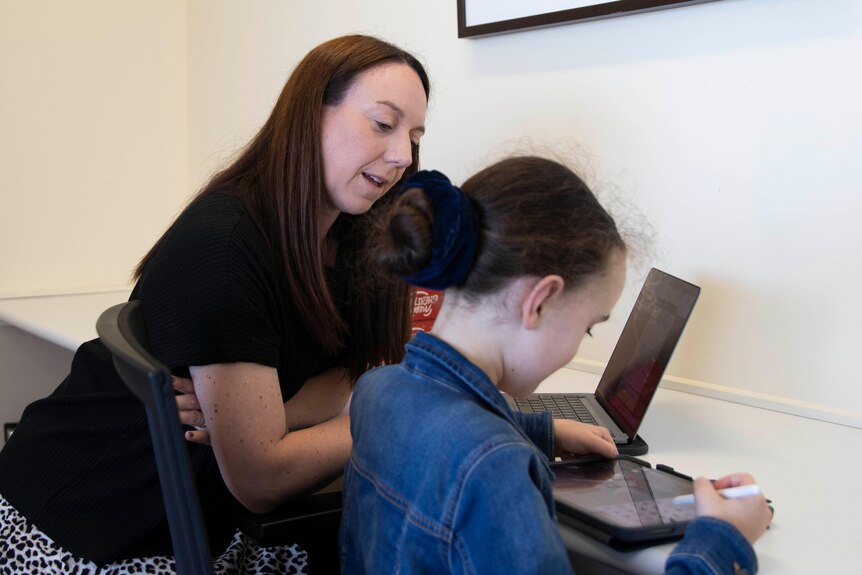 A woman with long brown hair talks to a child looking at an iPad