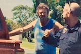 Gene Bristow, in a singlet, leans on a car as he speaks to police.