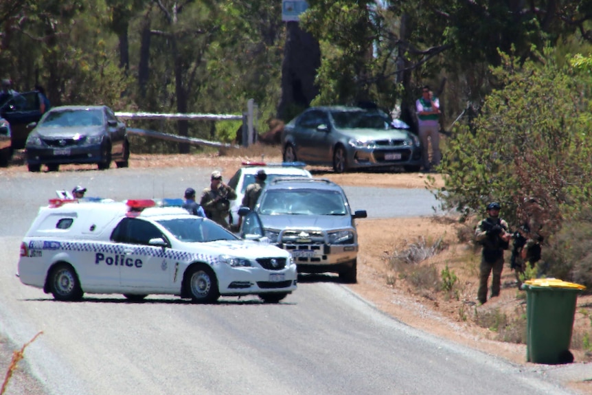 A wide shot with police officers and other emergency services workers and several police cars at the end of a semi-rural road.