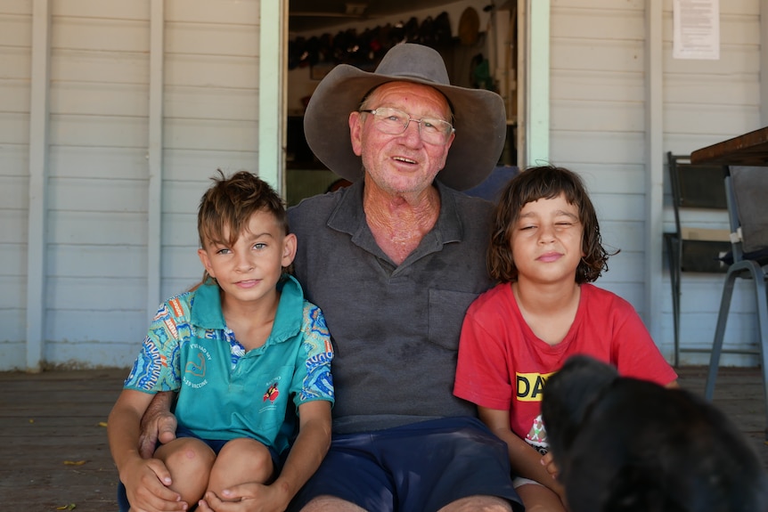 An older man wearing a cowboy hat sits on a wooden porch with arms around two small boys