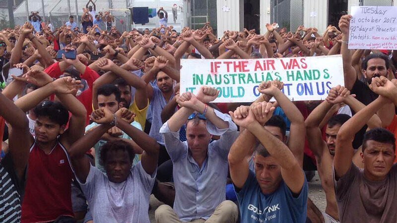 A large group of people sitting on the ground with their arms raised above their heads as if handcuffed.