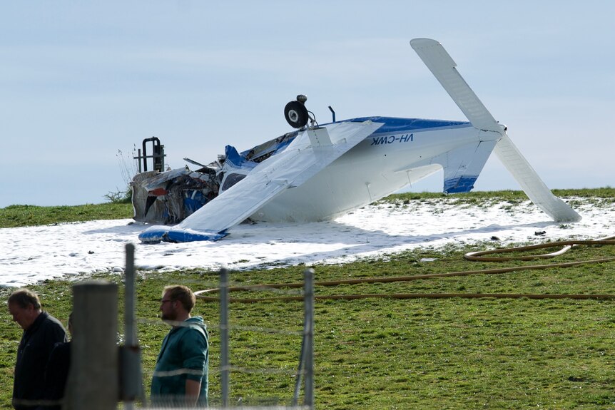 A white plane is crashed upside down on a green field, surrounded by white fire extinguisher foam.