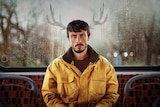 Donny stares into the camera as he sits at the back of a bus wearing a yellow jacket, with antlers photoshopped behind him.