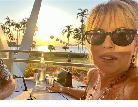 Blonde woman in glasses taking selfie in front of water front beach and palm trees