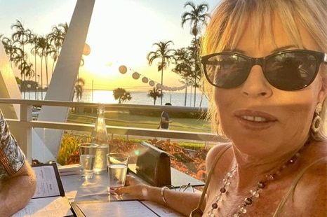 Blonde woman in glasses taking selfie in front of water front beach and palm trees