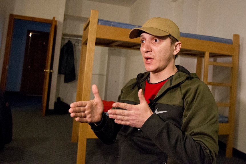 A harrowed looking man wearing a cap in a room next to a bunk bed gestures while speaking.