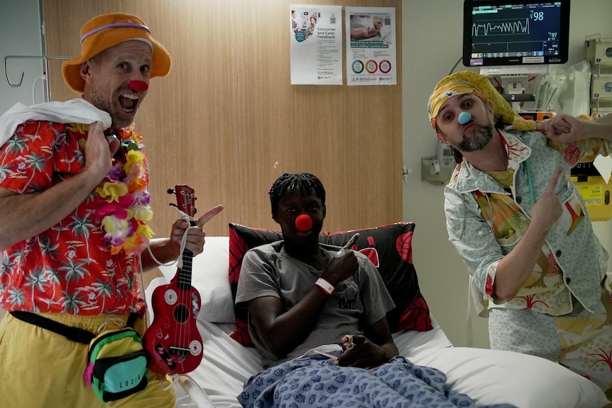 Sekou smiles, wearing a red nose, with the two clown doctors standing on either side of his hospital bed.